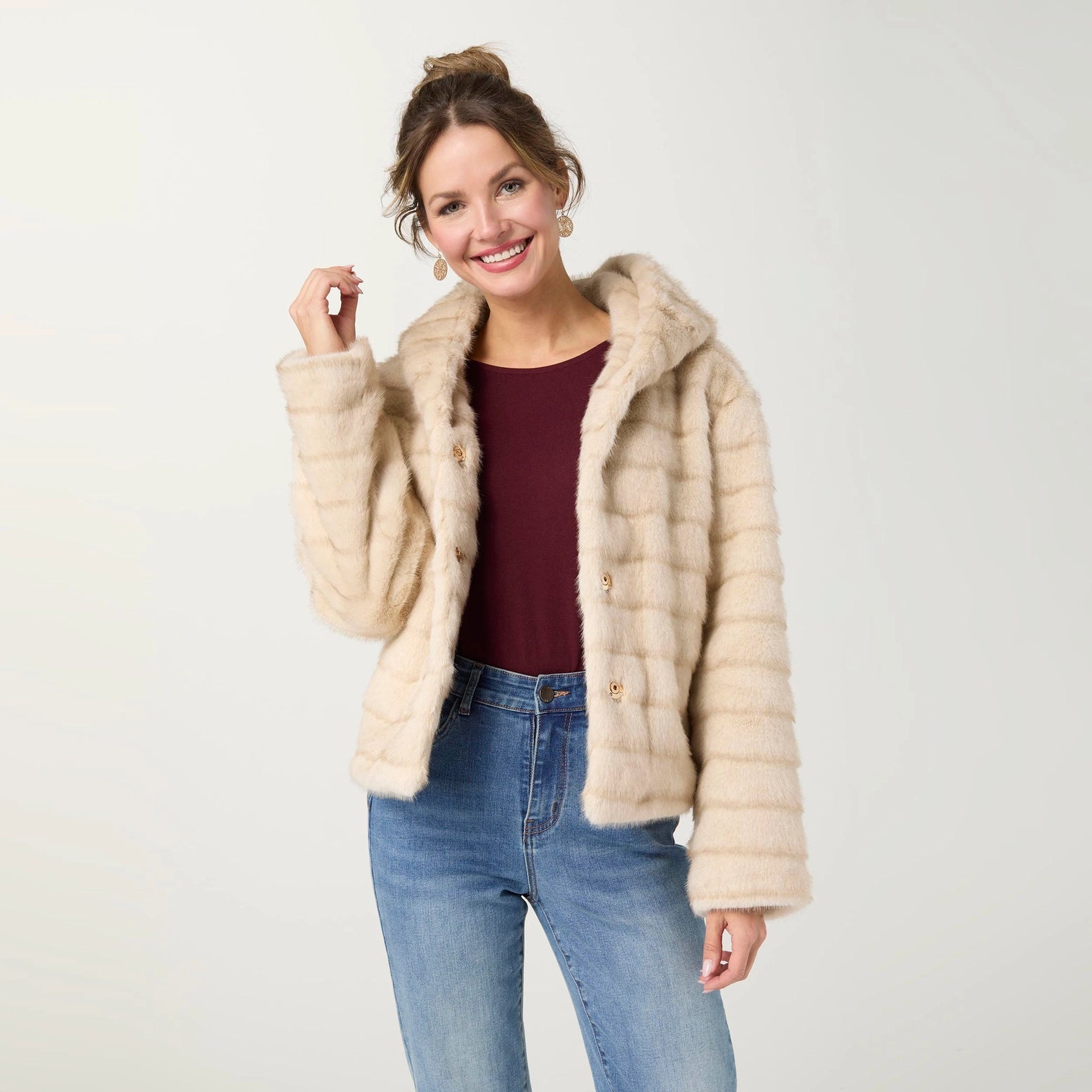 A woman modeling a light beige faux fur jacket with a hood, worn over a burgundy top and blue jeans, against a plain light gray background.