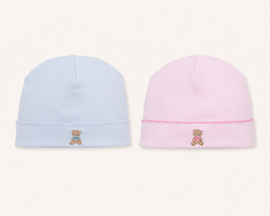 Two babykissy kissy teddy bear club beanie hats on a light cream background, one light blue with a small embroidered teddy bear and the other light pink with a matching pink embroidered teddy bear and decorative pink stitched trim.