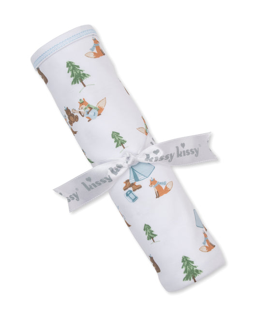 A rolled white baby blanket with a woodland print of foxes, trees, and a tent, tied with a white ribbon that reads 'kissy kissy'.