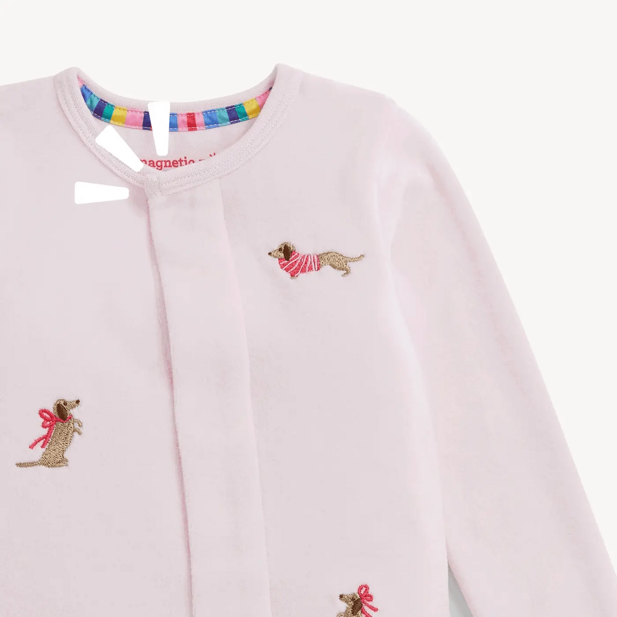 A close-up of a light pink baby velour footie featuring embroidered dachshund dogs wearing red-and-white striped sweaters and small red bows. The cardigan has a round neckline with a colorful striped inner trim.