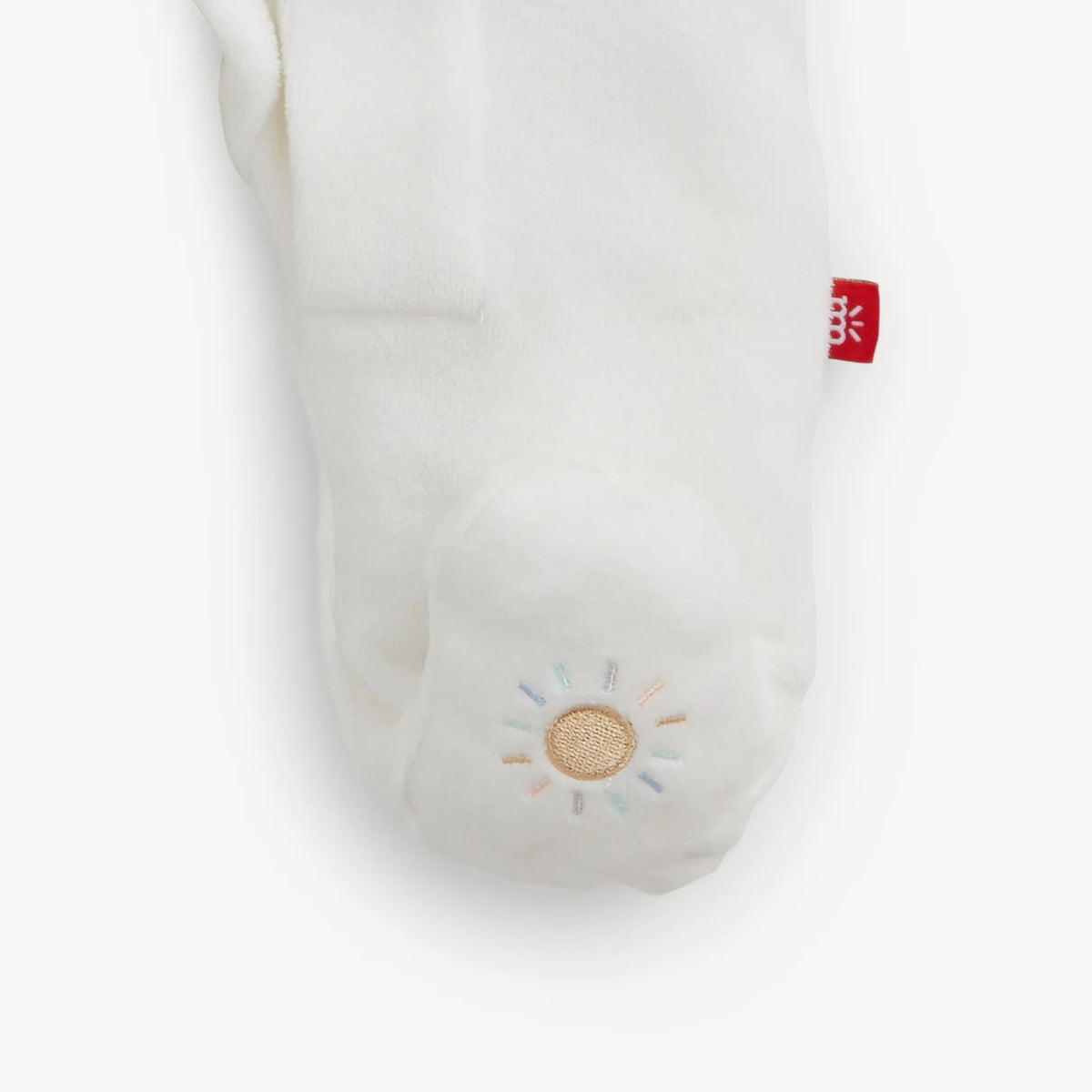 A close-up of a white plush footie with a small embroidered sun motif on the foot and a small red fabric tag near the side.