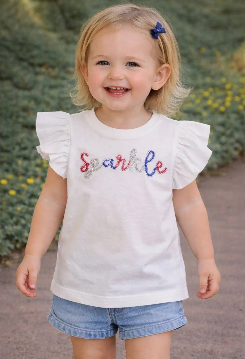 Child wearing a mudpie white shirt with 'Sparkle' in red white and blue tinsel  letters, standing outdoors.