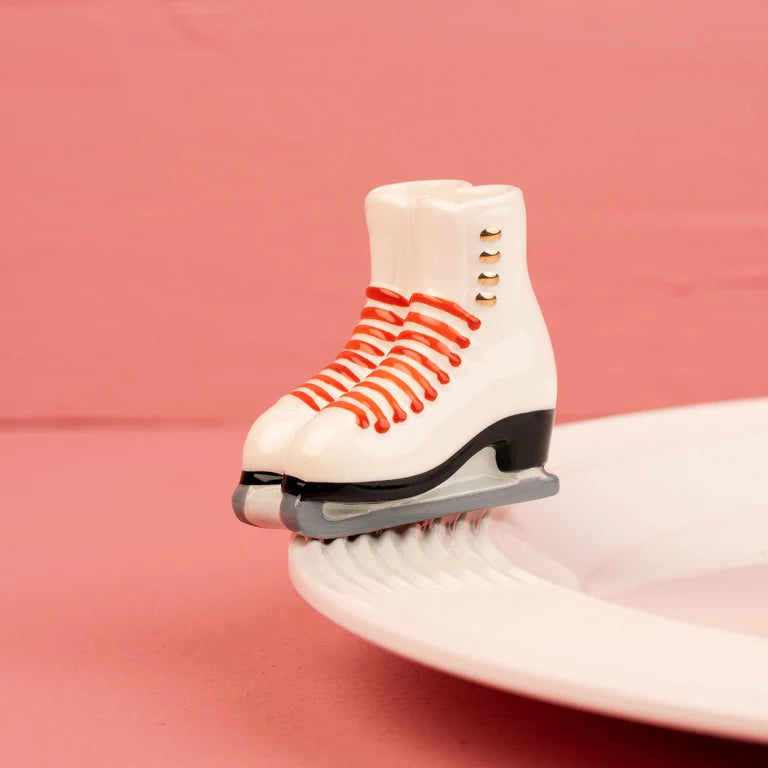 A miniature white ice skate Nora Fleming mini with red laces and a black blade, balanced on the edge of a white plate against a pink background.