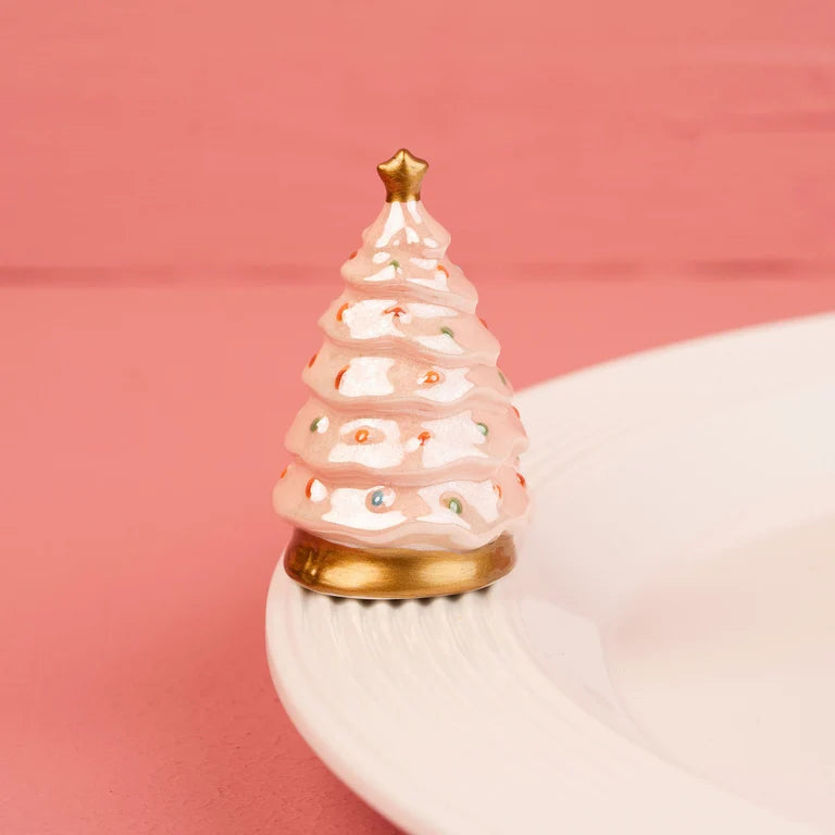A small decorative Christmas tree Nora Fleming mini with a pink and white frosting-like textured body, gold star topper, and gold base, displayed on a white plate against a pink background.