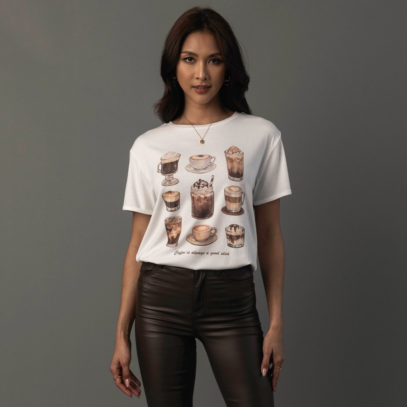 A woman modeling a white short-sleeve graphic t-shirt with a coffee-themed illustration print on the front, paired with dark brown faux-leather pants, against a solid dark gray studio background.