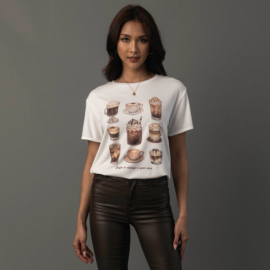 A woman modeling a white short-sleeve graphic t-shirt with a coffee-themed illustration print on the front, paired with dark brown faux-leather pants, against a solid dark gray studio background.