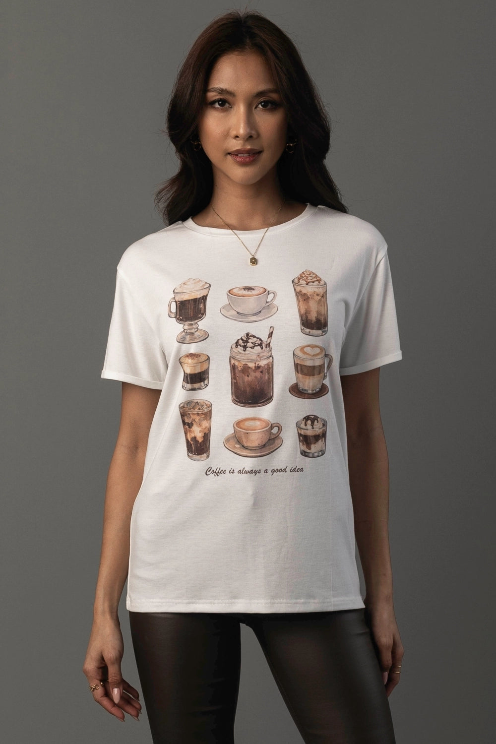 A woman modeling a white short-sleeve graphic t-shirt featuring illustrations of various coffee drinks and the text 'Coffee is always a good idea' on a solid gray background.