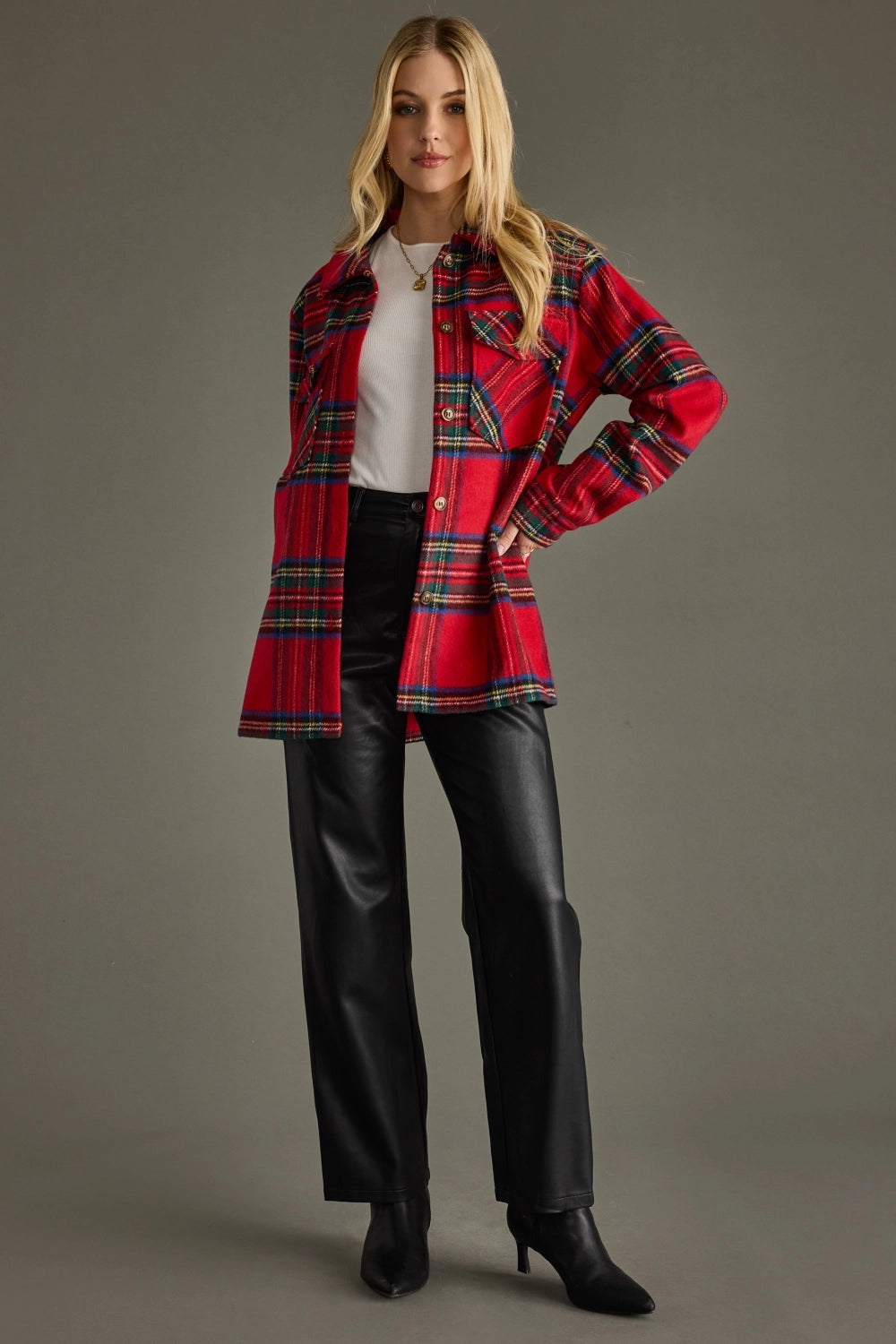 A model stands against a solid gray studio background wearing a red tartan plaid jacket over a white top, black wide-leg pants, and black heeled ankle boots.