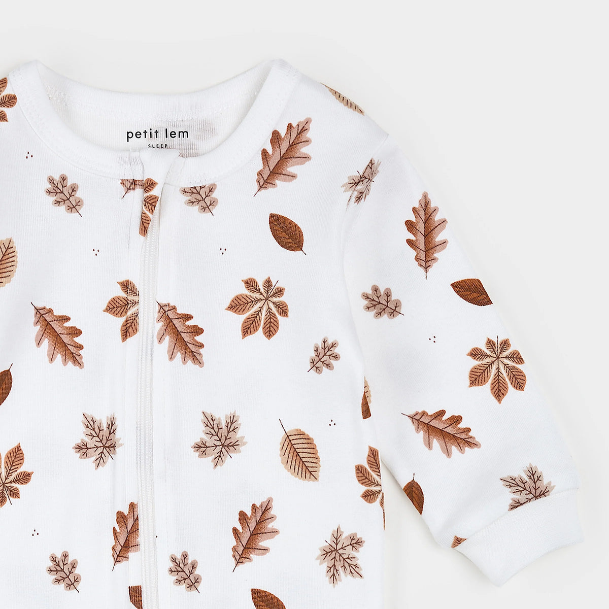 A close-up of a white baby zip-up sleeper with an all-over brown autumn leaf pattern, featuring the brand name 'petit lem' printed inside the collar.