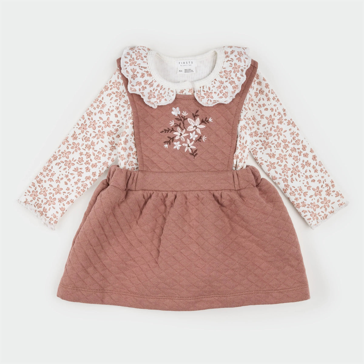 A baby girl's long-sleeve dress featuring a dusty rose quilted pinafore bodice with embroidered white flowers, layered over a white long-sleeve shirt with a dusty rose floral print and a white lace ruffled collar.