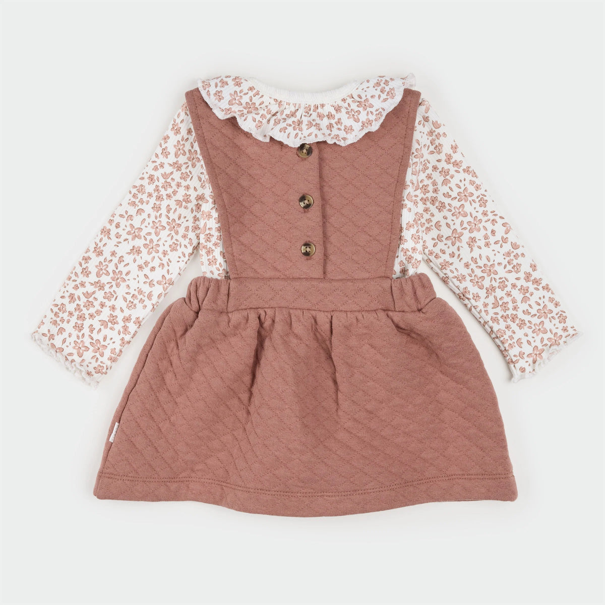 A baby girl's long-sleeve dress featuring a dusty rose quilted pinafore bodice with three buttons, layered over a white long-sleeve shirt with a dusty rose floral print and a white lace ruffled collar.
