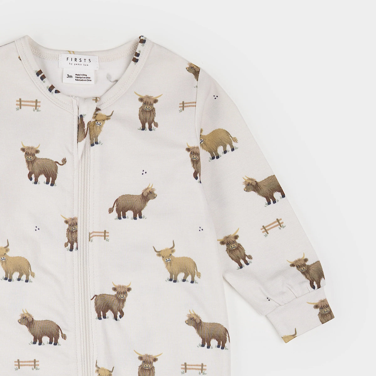 A long-sleeve baby zip-up sleeper, featuring an all-over print of Highland cows and small wooden fences on a warm off-white background.