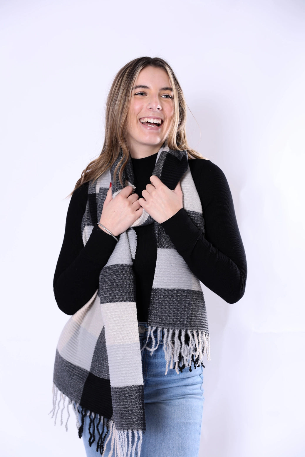 A woman smiling and wearing a black, gray, and white plaid fringed scarf over a black long-sleeve top and blue jeans, photographed against a plain white studio background.