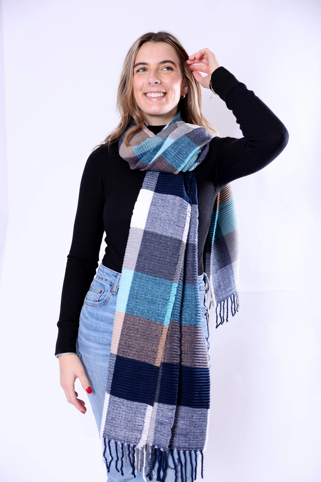 A woman modeling a large plaid scarf with blue, gray, beige, and white blocks, paired with a black long-sleeve top and light blue jeans against a white studio background.