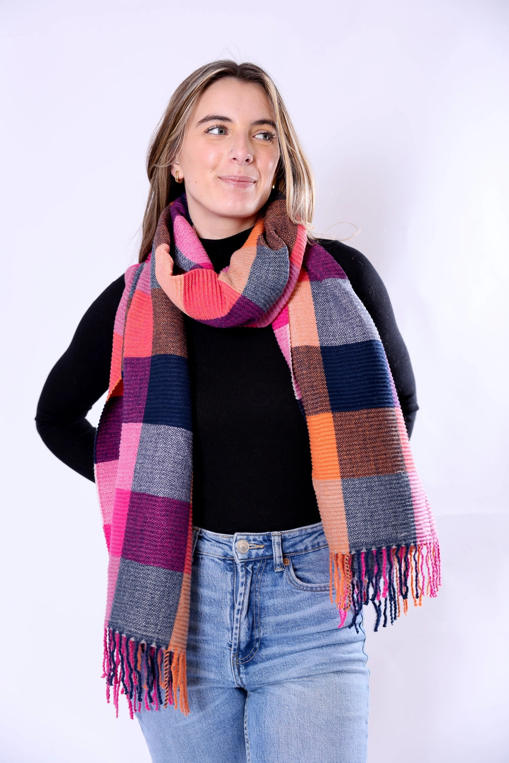 A woman wearing a large multicolor plaid scarf with pink, orange, purple, blue, gray, and brown blocks, styled over a black top and blue jeans against a plain white studio background.