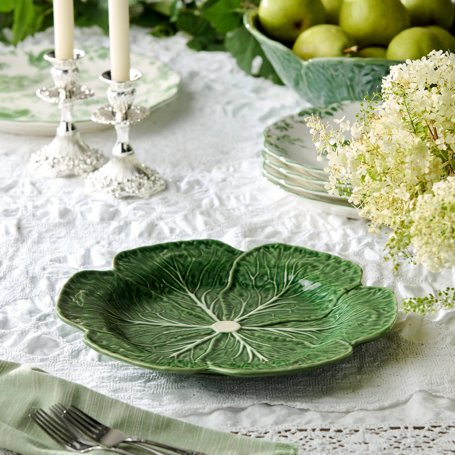 Green leaf-shaped plate on a table with white lace tablecloth, silver candlesticks, and green apples.