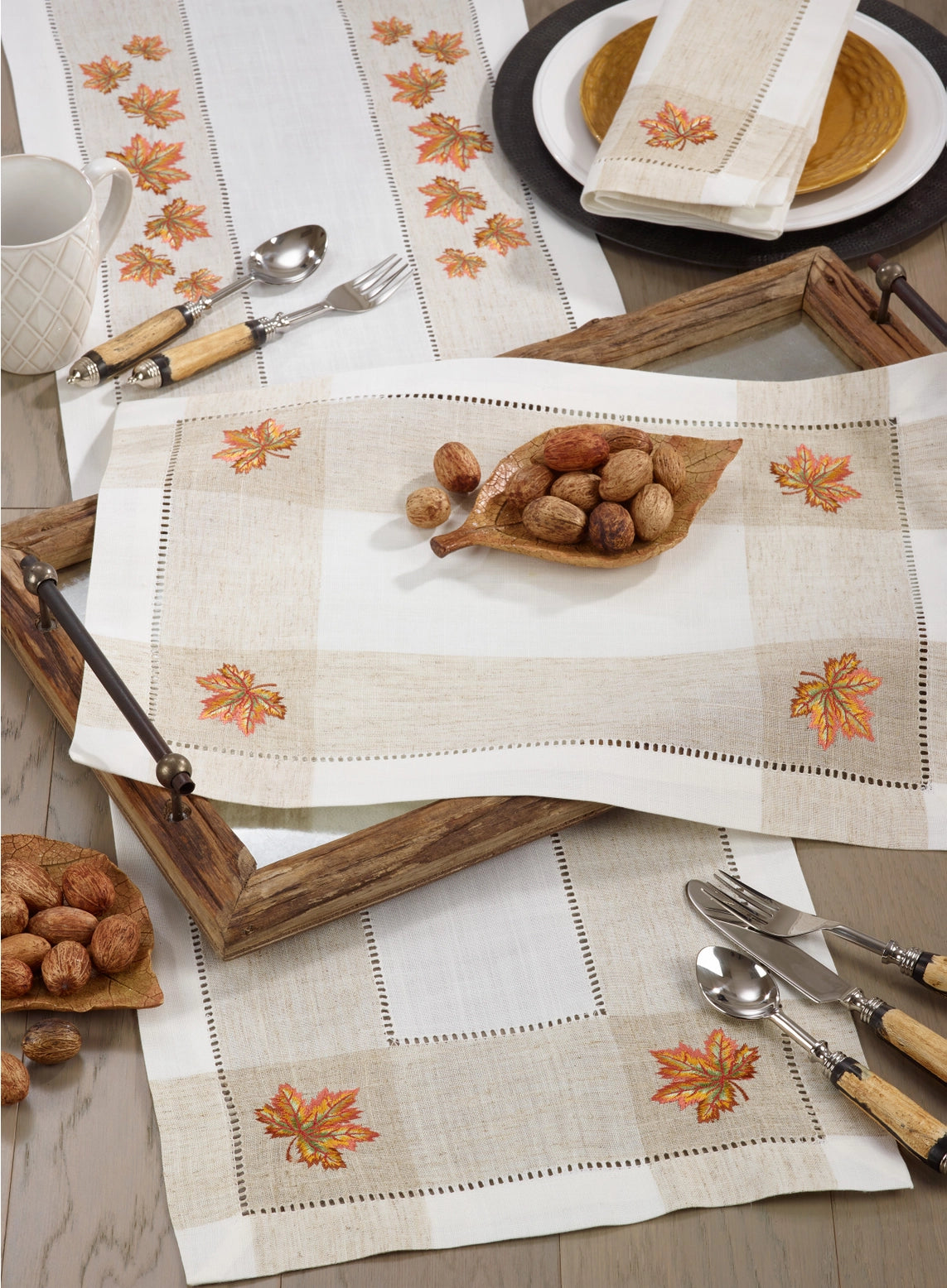 A styled tabletop setting featuring embroidered checkered table linens in a warm autumn color palette. The linens are off-white with beige gingham checks, orange and brown embroidered maple leaves, and delicate dotted borders.