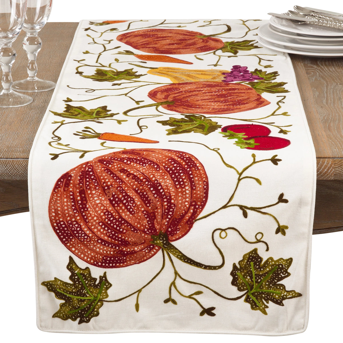 An embroidered table runner with a white base featuring large orange pumpkins, green leaves, carrots, and gourds, styled on a wooden dining table with wine glasses and plates.