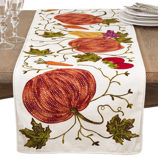An embroidered table runner with a white base featuring large orange pumpkins, green leaves, carrots, and gourds, styled on a wooden dining table with wine glasses and plates.