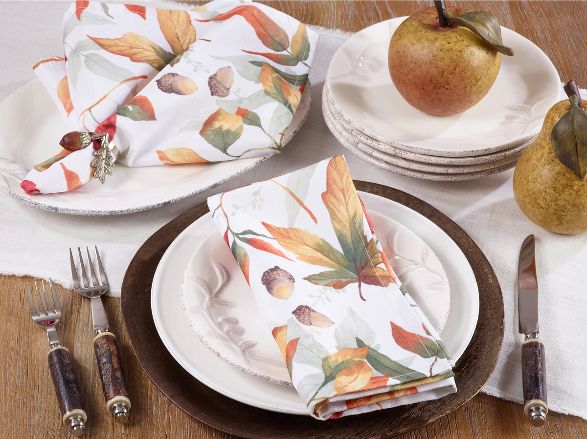 An autumn-themed table setting featuring white napkins with a printed pattern of orange, yellow, and green leaves and brown acorns, styled on white plates with dark brown chargers, silverware, and decorative pears on a wooden table.