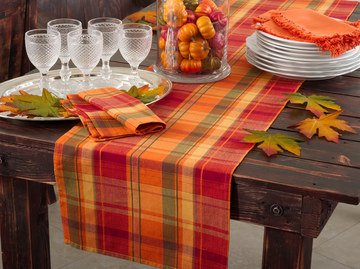 An autumn-themed table runner with a red, orange, and yellow plaid pattern draped over a dark wooden dining table, styled with matching napkins, white plates, wine glasses, and fall leaves.