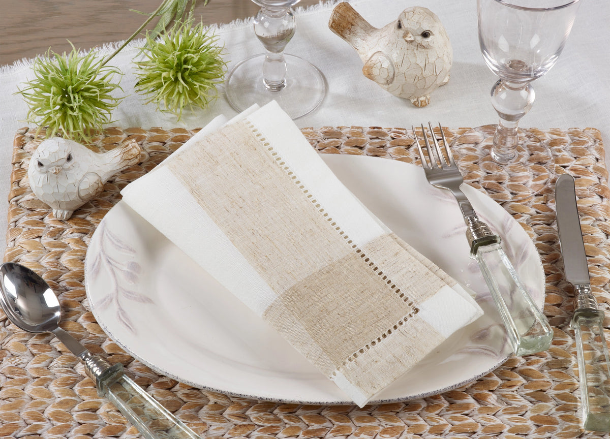 A place setting featuring a folded beige and white woven napkin on a white plate, surrounded by clear glassware, silver cutlery, and decorative bird figurines on a woven placemat.