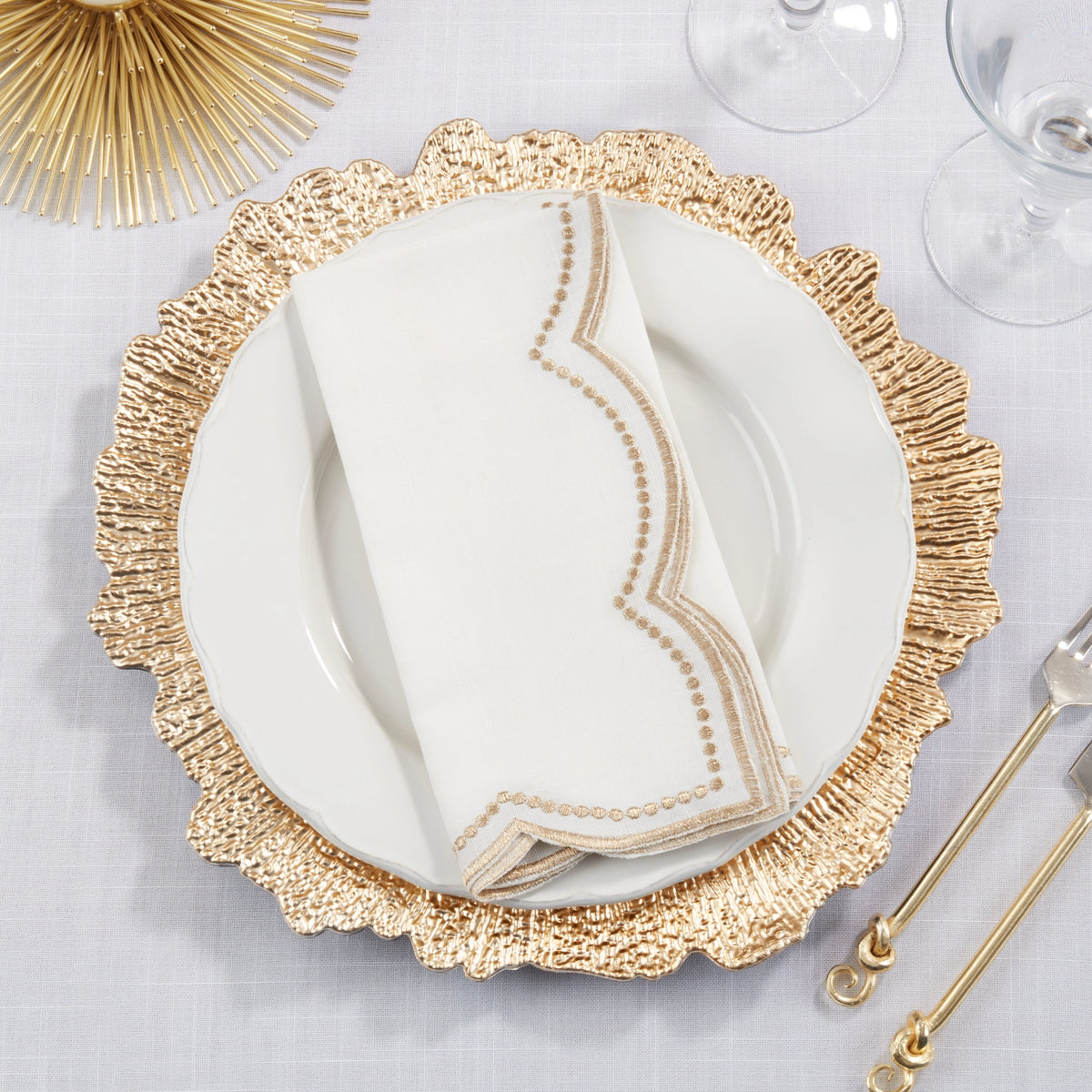 Gold-rimmed plate with a white napkin with scallop edge on a gray surface, with gold cutlery and decorative elements.