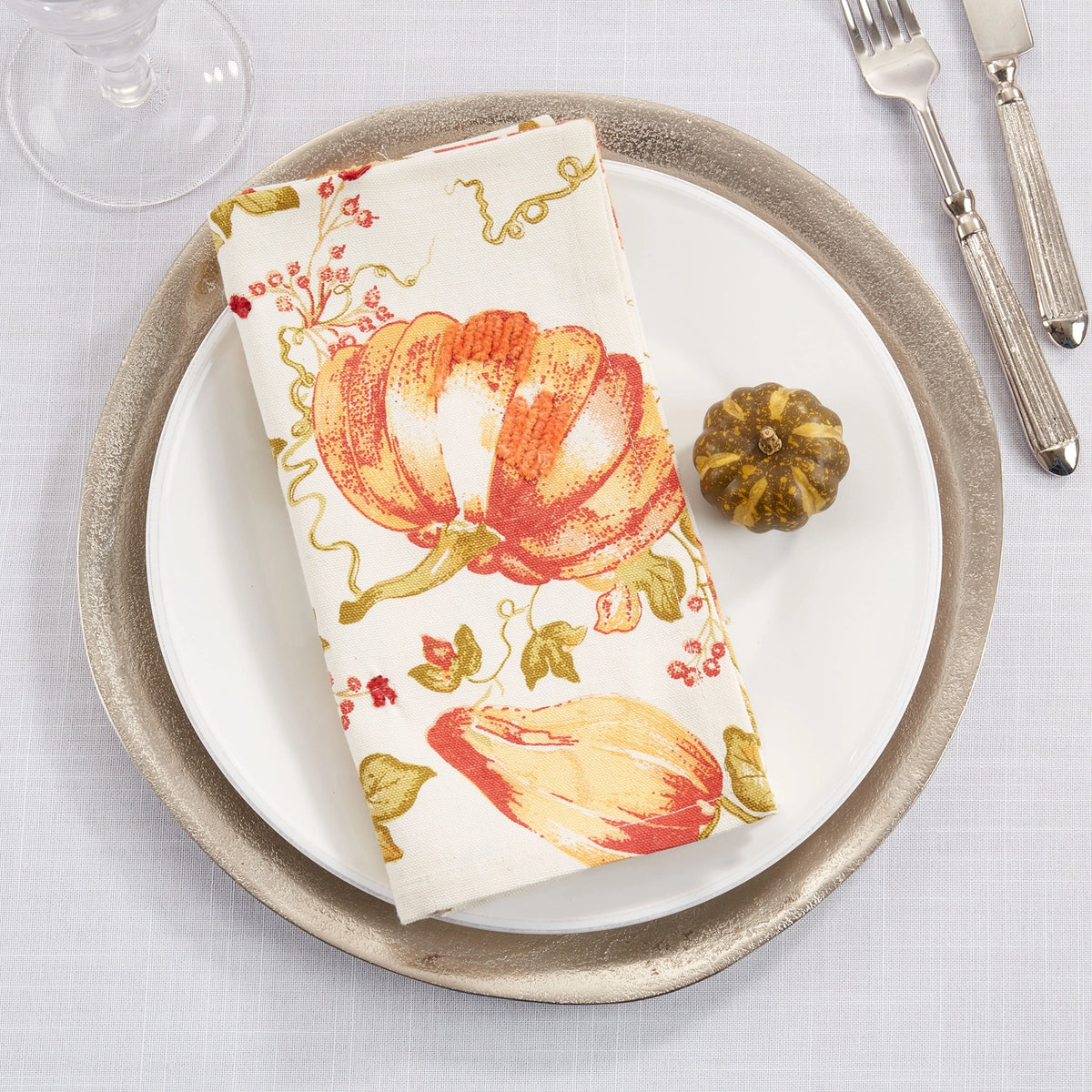 A folded white napkin with a red, orange, and green autumn pumpkin print, styled on a white plate with a rustic beige charger plate, a small decorative pumpkin, and silverware on a light tablecloth.