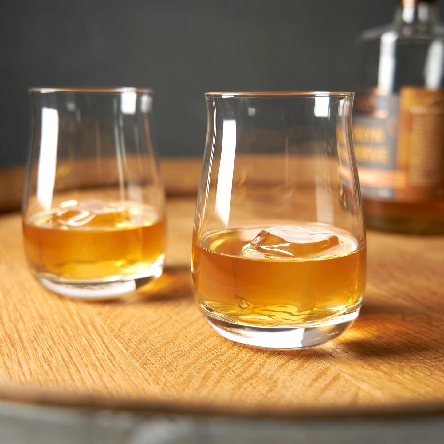 Two spiegalau glasses of whiskey with a bottle in the background on a wooden surface.