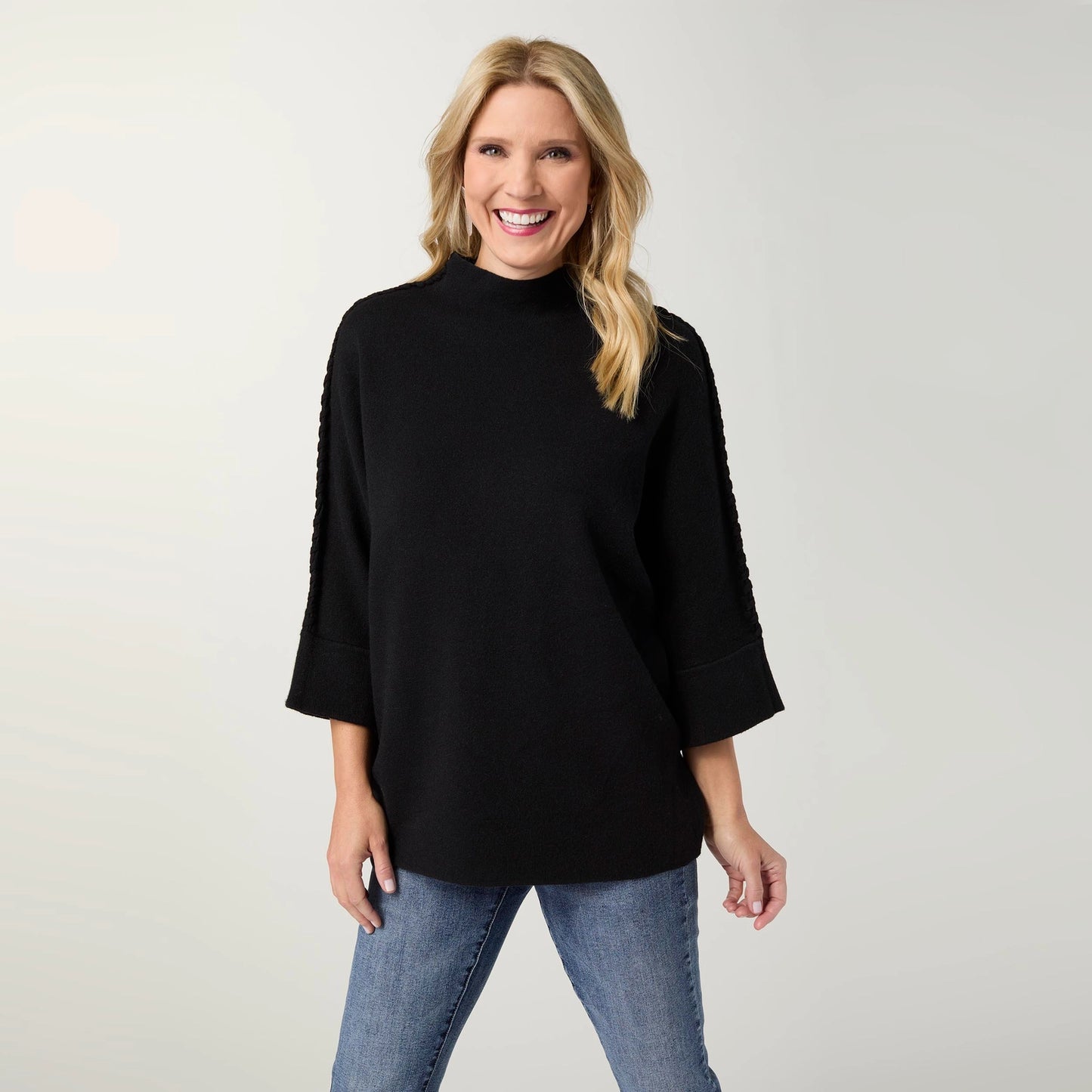 A woman wearing a black mock-neck sweater with textured sleeve detailing and blue jeans, standing against a light gray studio background.