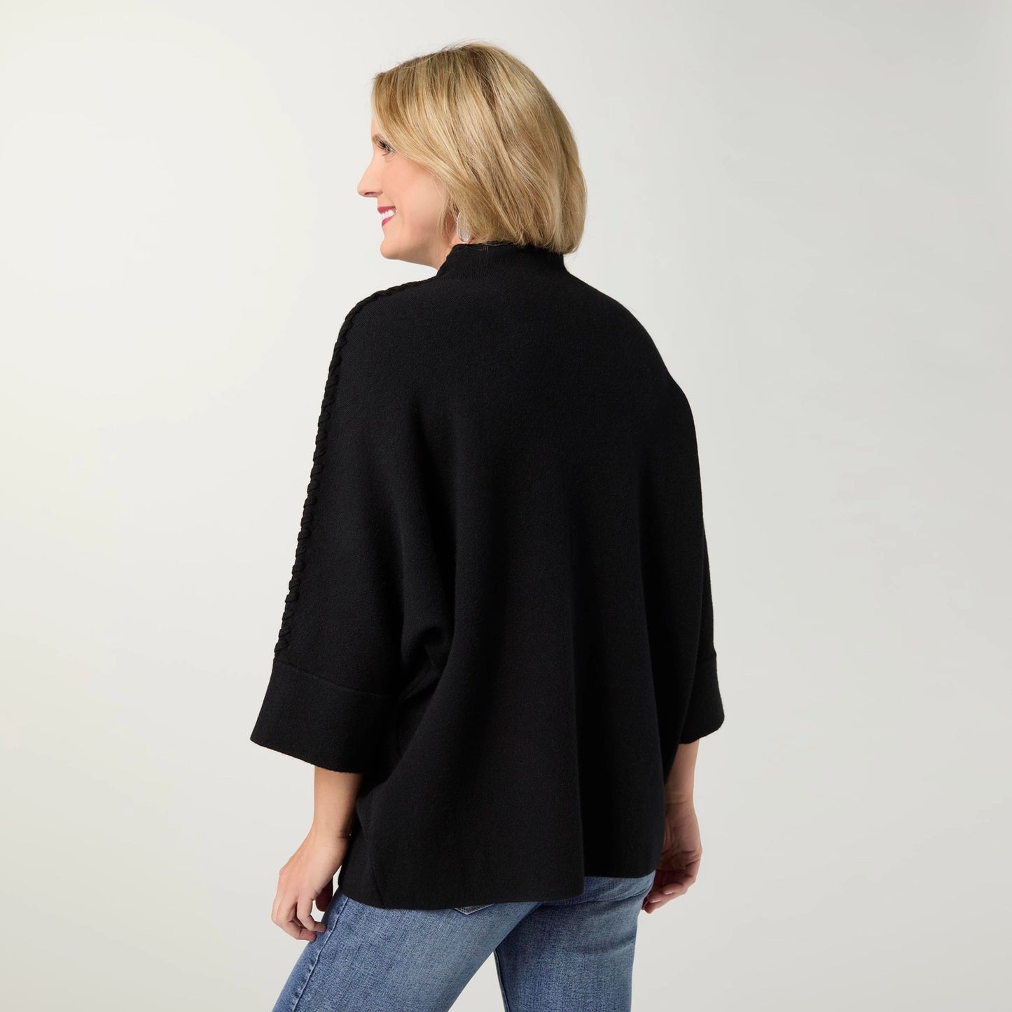A woman is shown from the back wearing a black knit poncho-style top with textured sleeve detailing, paired with blue jeans, against a plain light gray studio background.