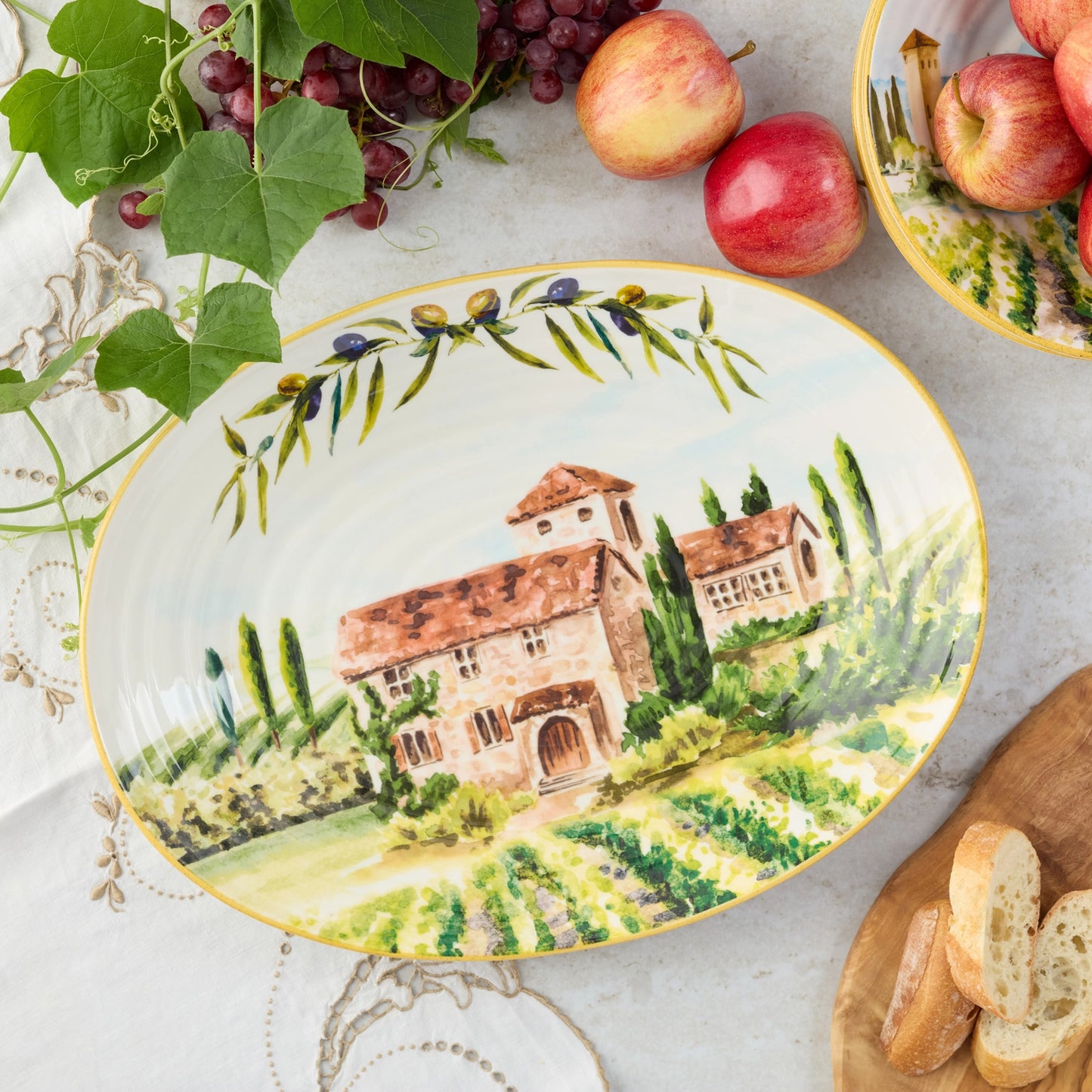 Decorative oval ceramic platter with a scenic tuscany design of houses and trees, placed on a table with apples and bread.