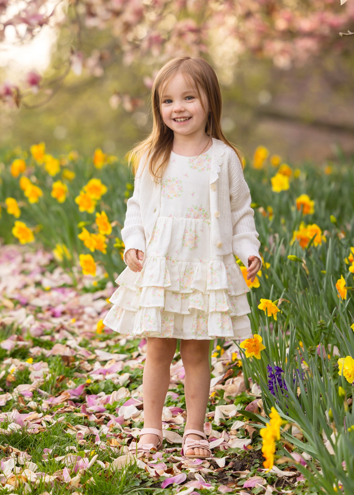 Young girl in a floral dress and a white Mabel and Honey Monica cardigan sweater standing in a field of flowers with cherry blossoms in the background