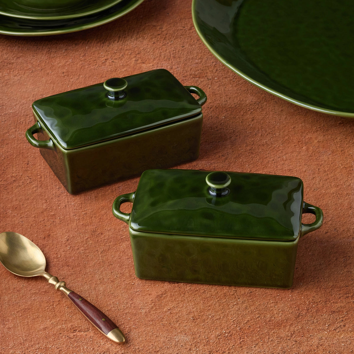 Two dark green glazed ceramic mini bakers with lids and side handles, arranged on a terracotta surface with a spoon and plates partially visible.